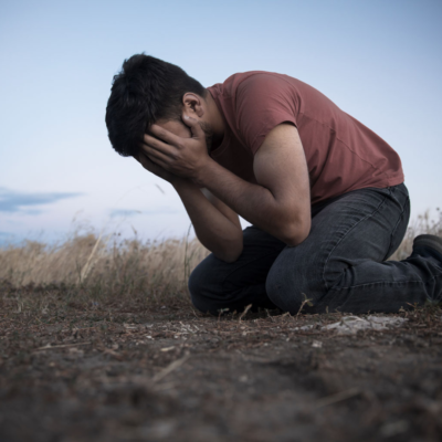 A person is sitting on the ground outdoors in distress. Their hands are covering their face, and they are leaning over toward the ground. 